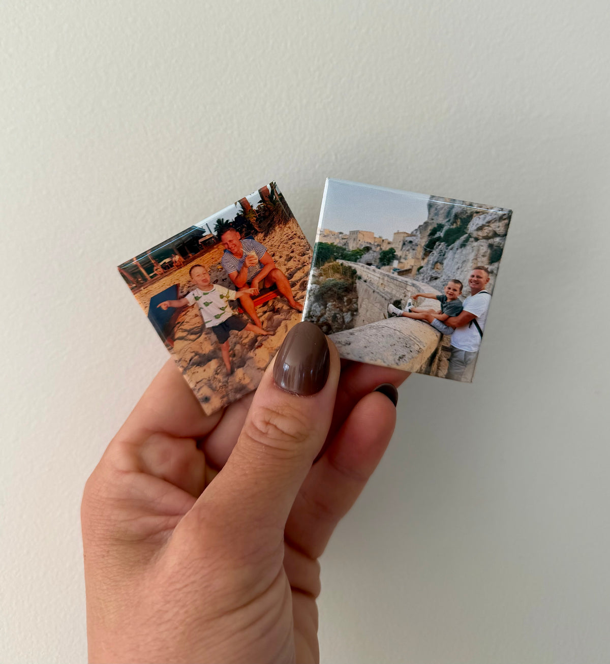A hand with brown nail polish holds two small square personalised photo magnets against a plain off-white wall. One magnet shows father and son in Italy, and the other shows father and son at the beach